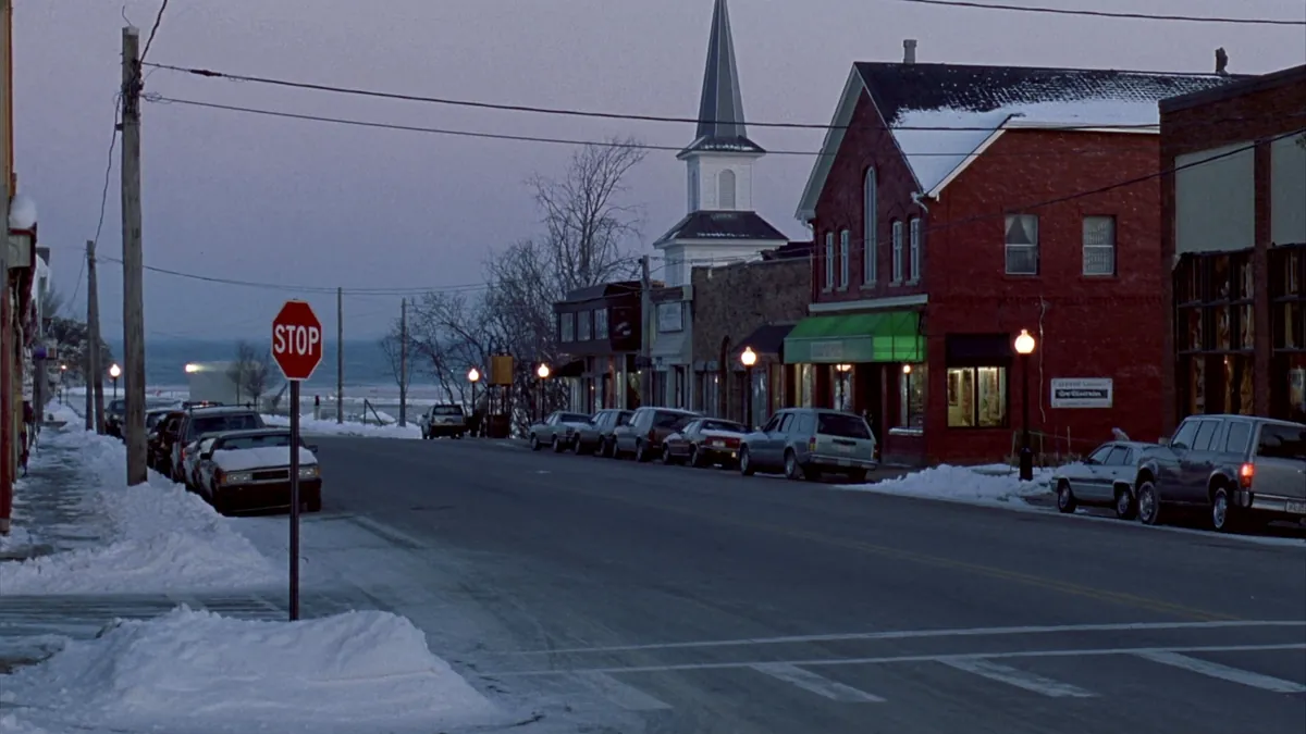 A snowy small-town main street in northern Michigan at dusk with brick storefronts, a church steeple, and Lake Michigan in the distance